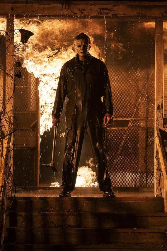 Eine maskierte Person im dunklen Overall steht vor einer brennenden Tür und hält ein Werkzeug.