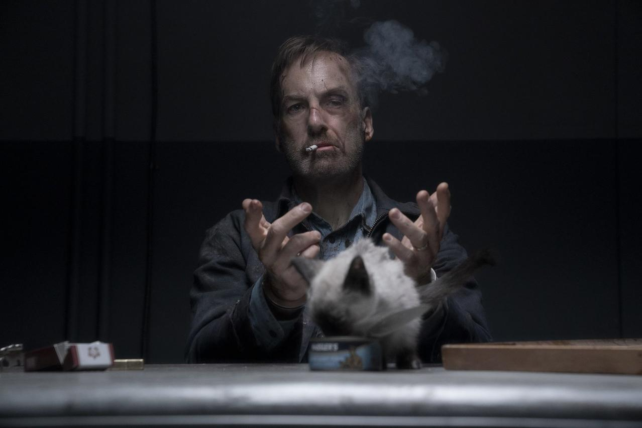 Ein Mann raucht, starrt vor sich hin und hält seine Hände hoch. Eine Katze frisst daneben.