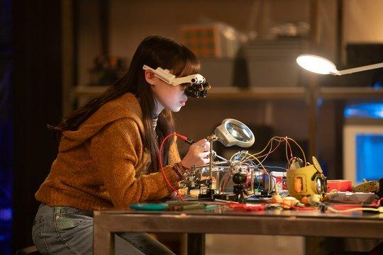 Ein Mädchen trägt eine Lupenbrille und arbeitet an einem komplexen Elektronikprojekt am Schreibtisch mit Lampe.