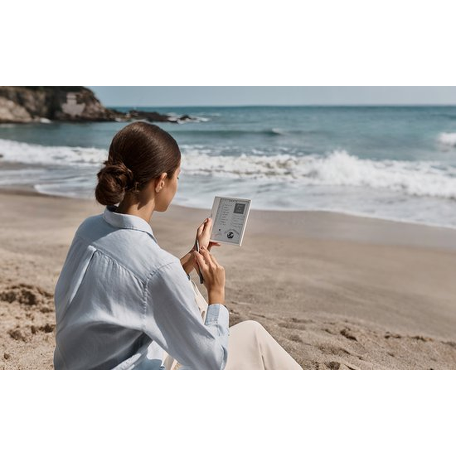 Vrouw met knot in haar leest e-reader op zandstrand, oceaan golven op achtergrond.