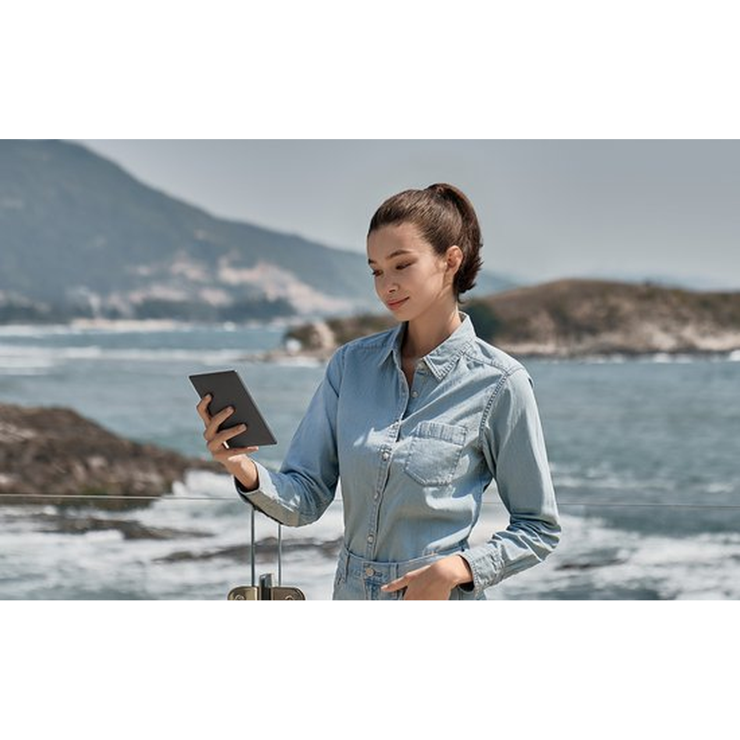 Vrouw in spijkerblouse met e-reader aan zee met bergen op achtergrond.