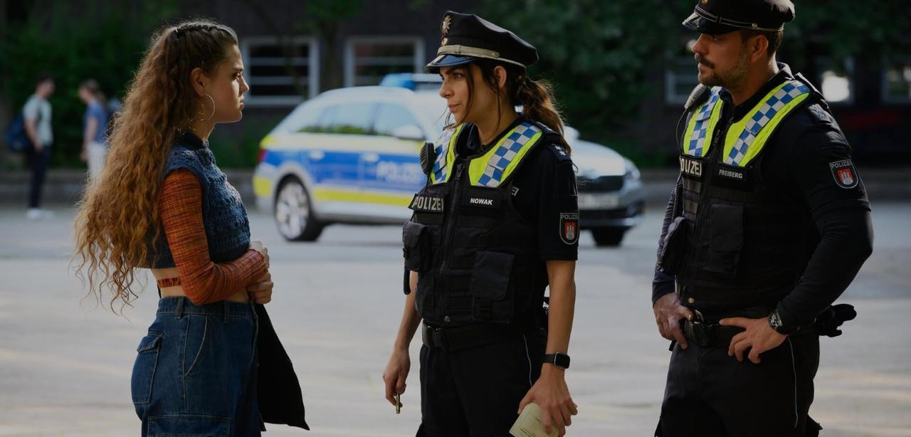 Zwei Polizisten sprechen mit einer jungen Frau vor einem Polizeiauto.