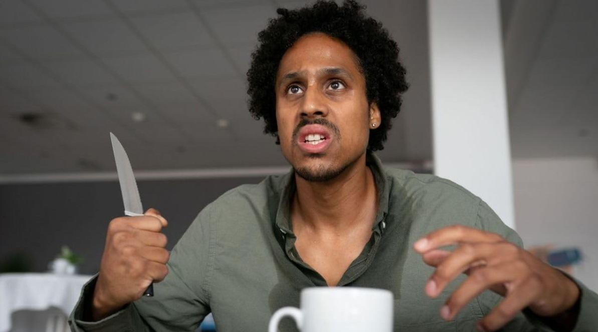 Mann mit Afro hält Messer, blickt intensiv nach oben. Weiße Tasse im Vordergrund.