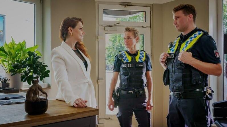 Eine Frau im weißen Blazer spricht mit zwei Polizisten in Uniform. Ein kleiner Baum steht auf einem Schreibtisch.