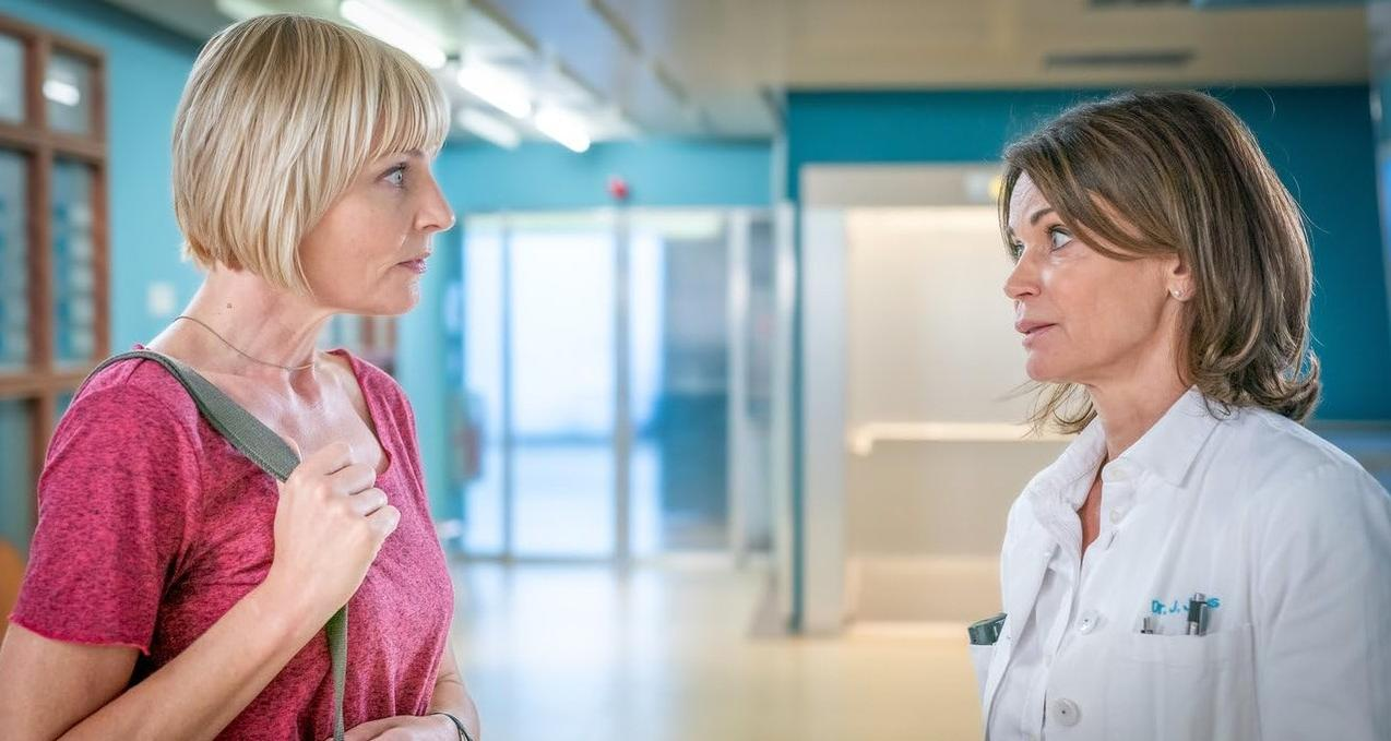 Zwei Frauen in einem Krankenhausflur. Eine trägt ein pinkes Hemd, die andere einen weißen Arztkittel.