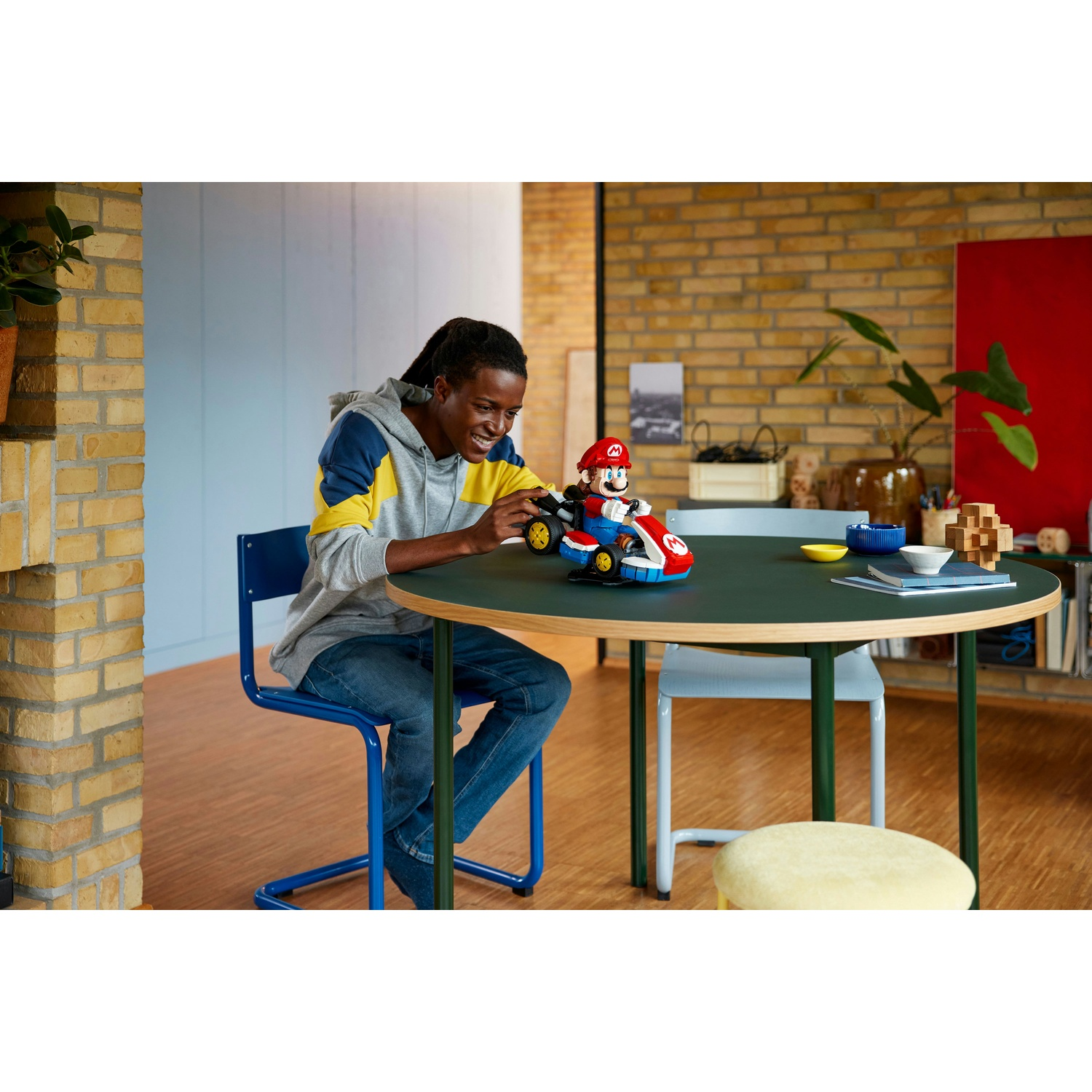 Una persona nera giovane è seduta a un tavolo rotondo, giocando con un set Lego di Mario Kart. Indossa una felpa grigia.