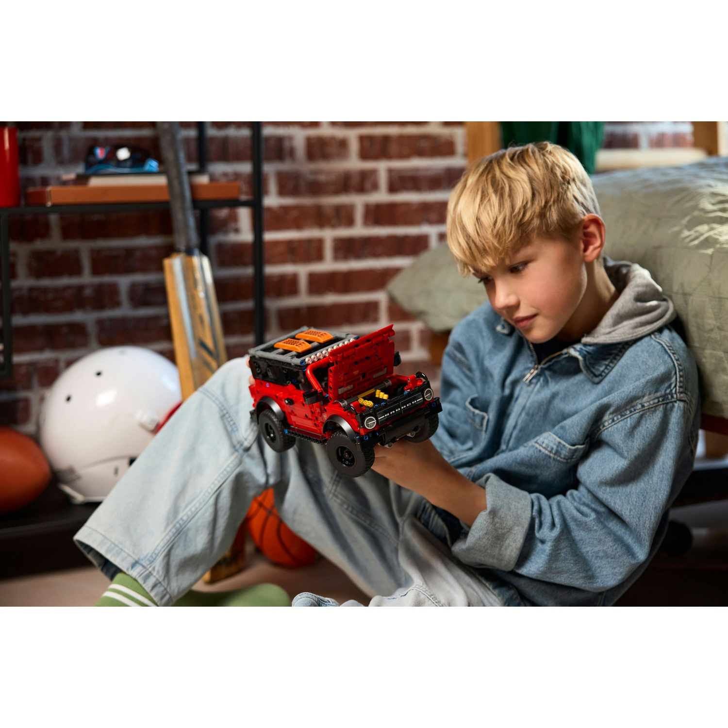 Ragazzo in giacca di jeans gioca con auto Lego rossa, circondato da attrezzature sportive.