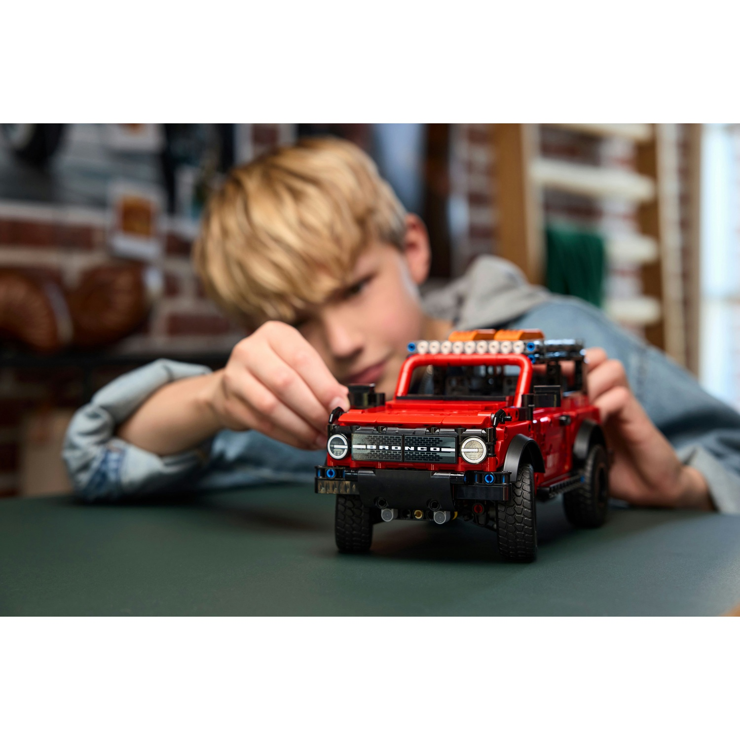 Un ragazzo in giacca di jeans monta un'auto modello Lego Ford Bronco rossa su un tavolo verde scuro.