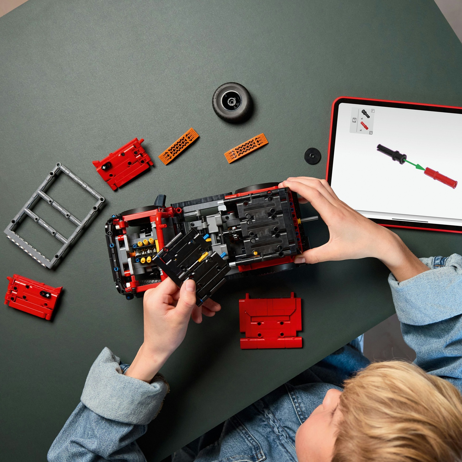 Bambino costruisce auto Lego rossa, segue istruzioni su tablet. Pezzi sul tavolo.
