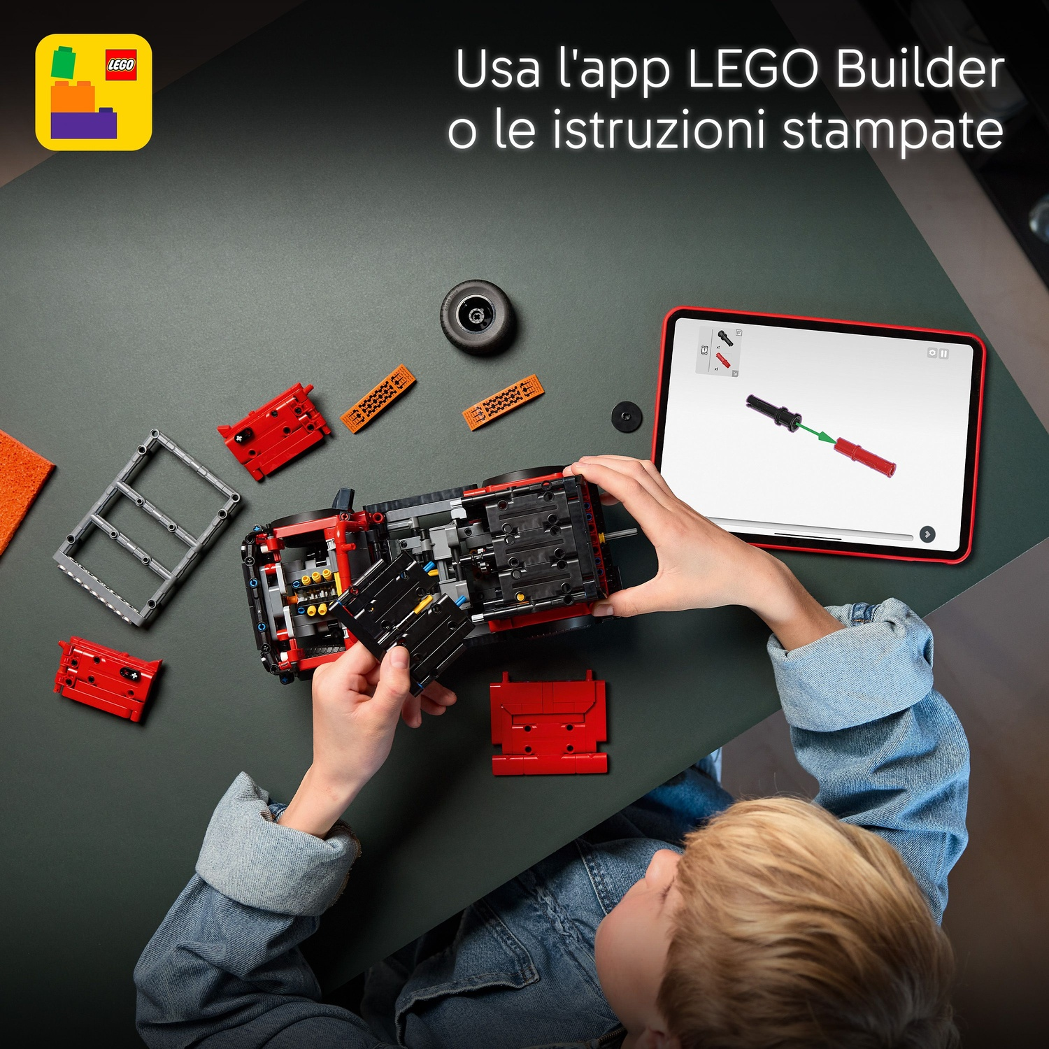Un bambino costruisce un'auto LEGO rossa seguendo istruzioni su tablet. Pezzi e attrezzi sparsi su un tavolo scuro.