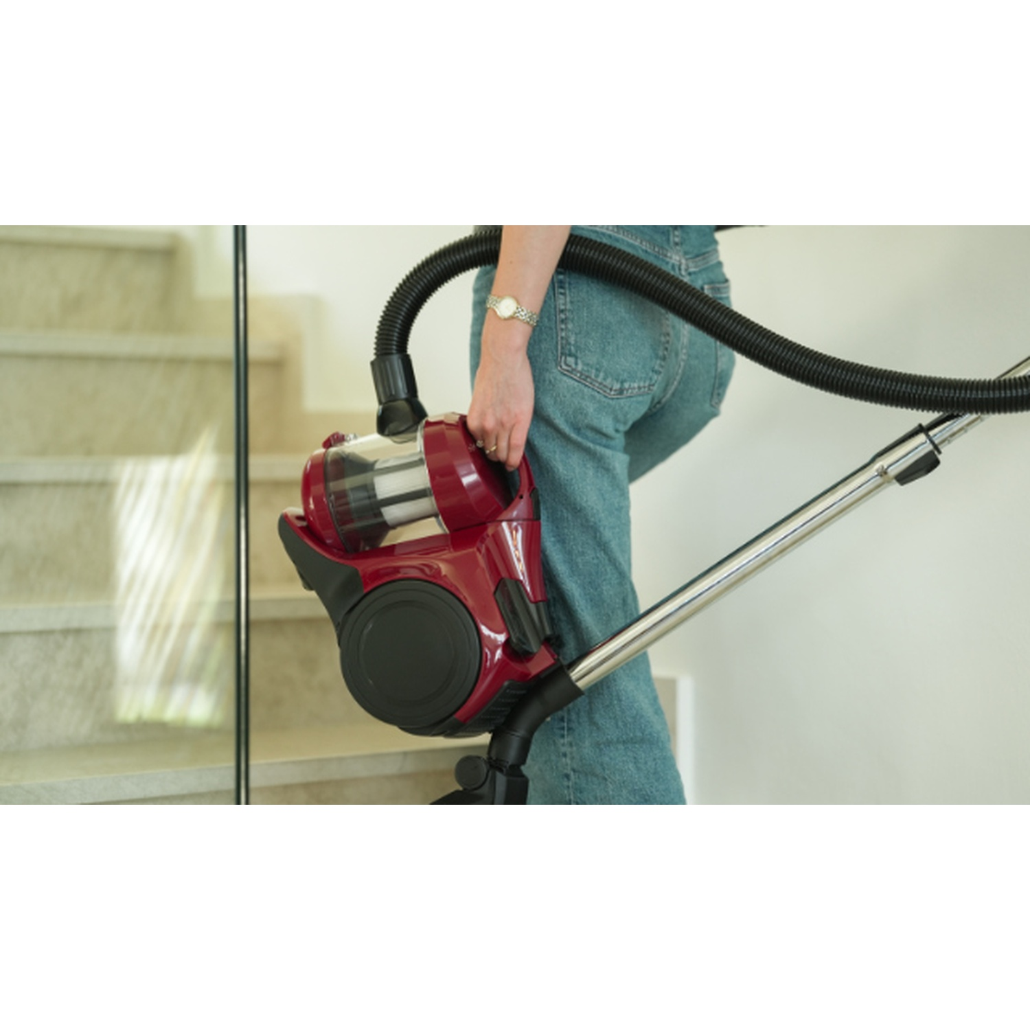 Persona in jeans porta aspirapolvere rosso con tubo, vicino alle scale.