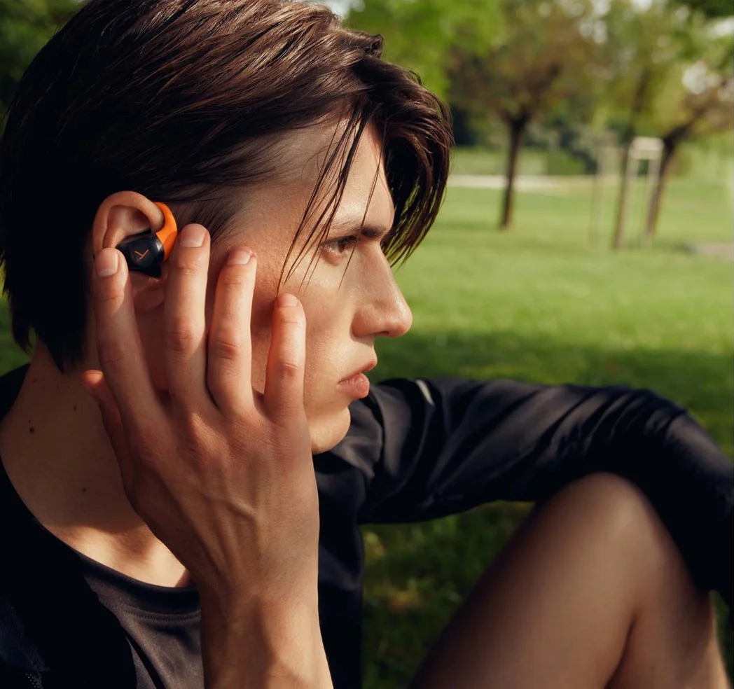 Homme en chemise noire avec écouteur orange dans un parc.