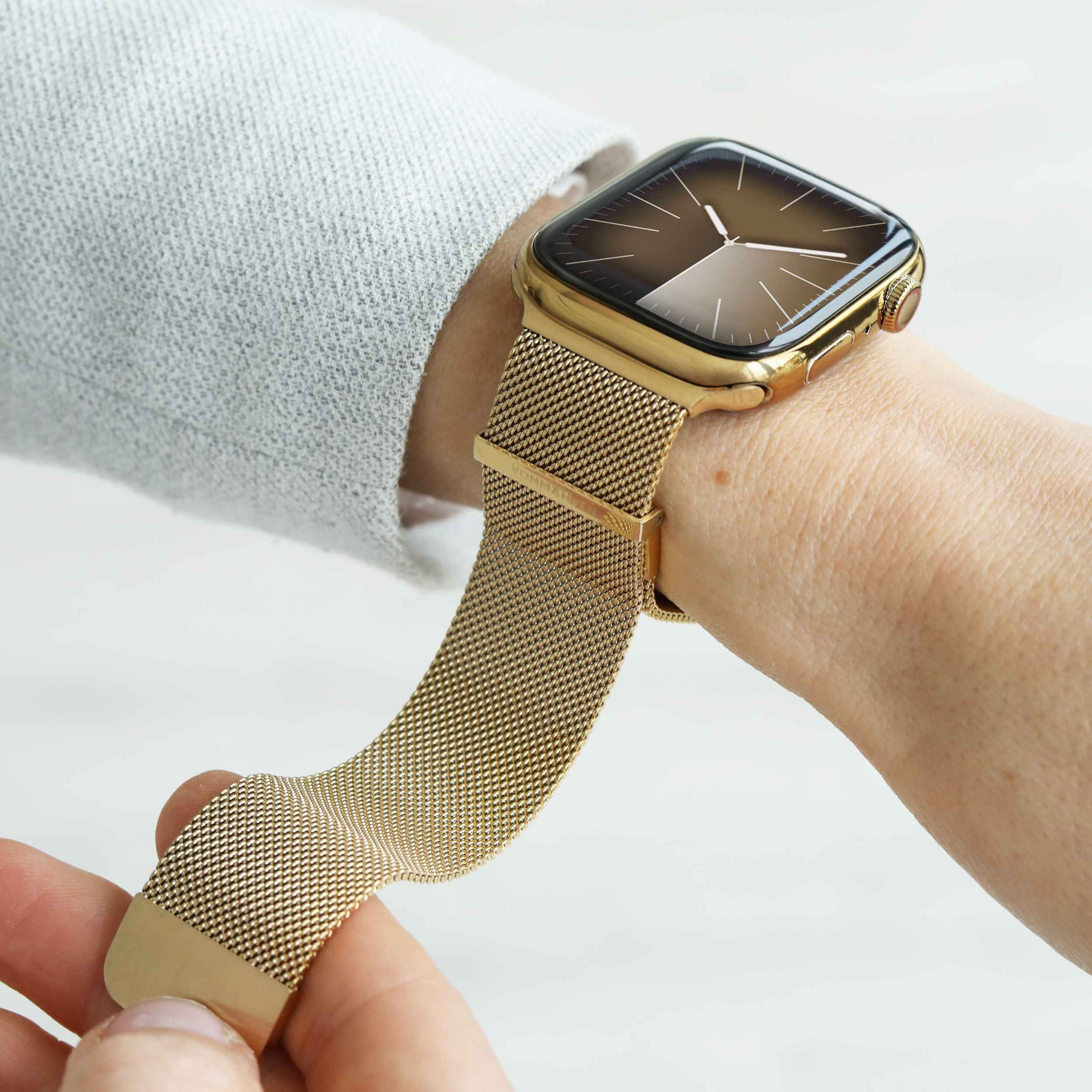 Une montre connectée dorée avec bracelet en maille sur un poignet couvert de tissu gris.
