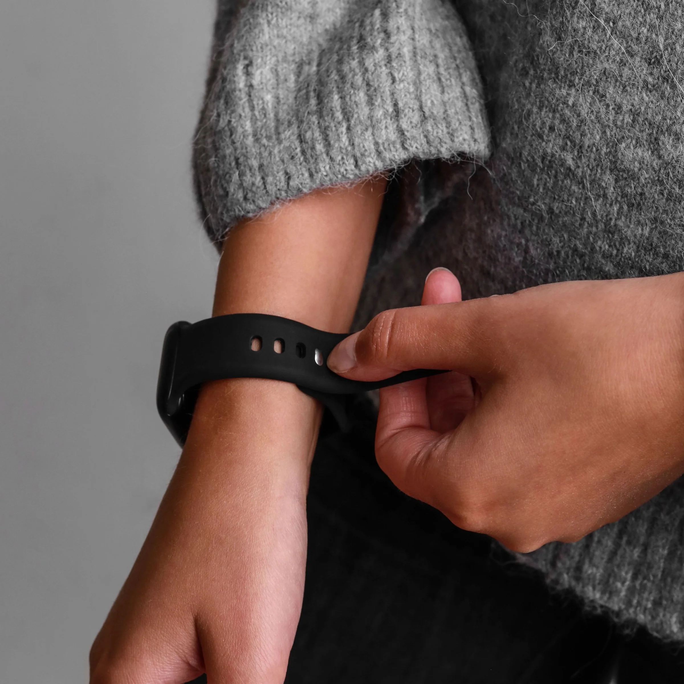 Eine Person in einem grauen Pullover befestigt ein schwarzes Uhrenarmband am Handgelenk.