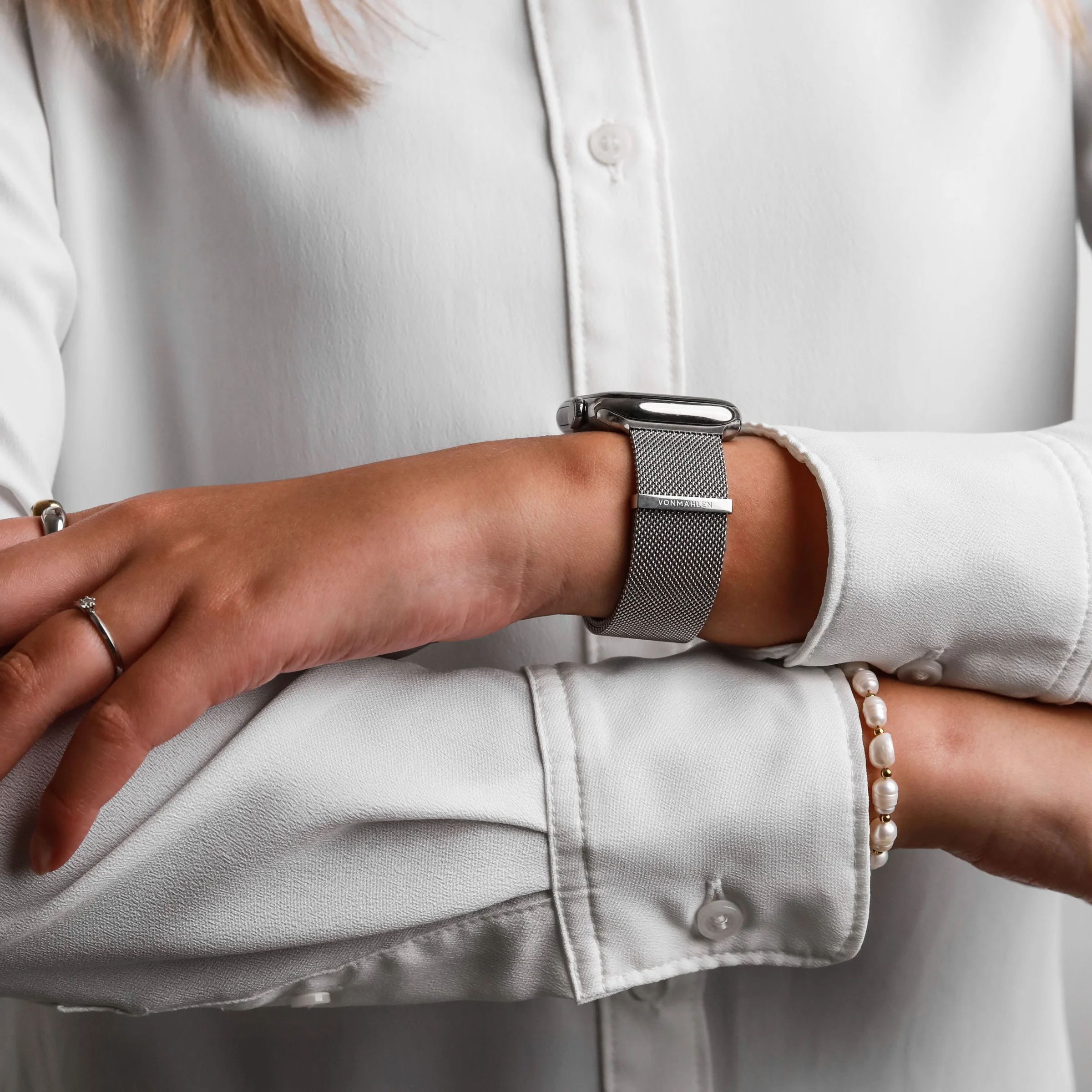 Une personne en chemise blanche porte une montre argentée en maille et un bracelet en perles. Bague diamant.