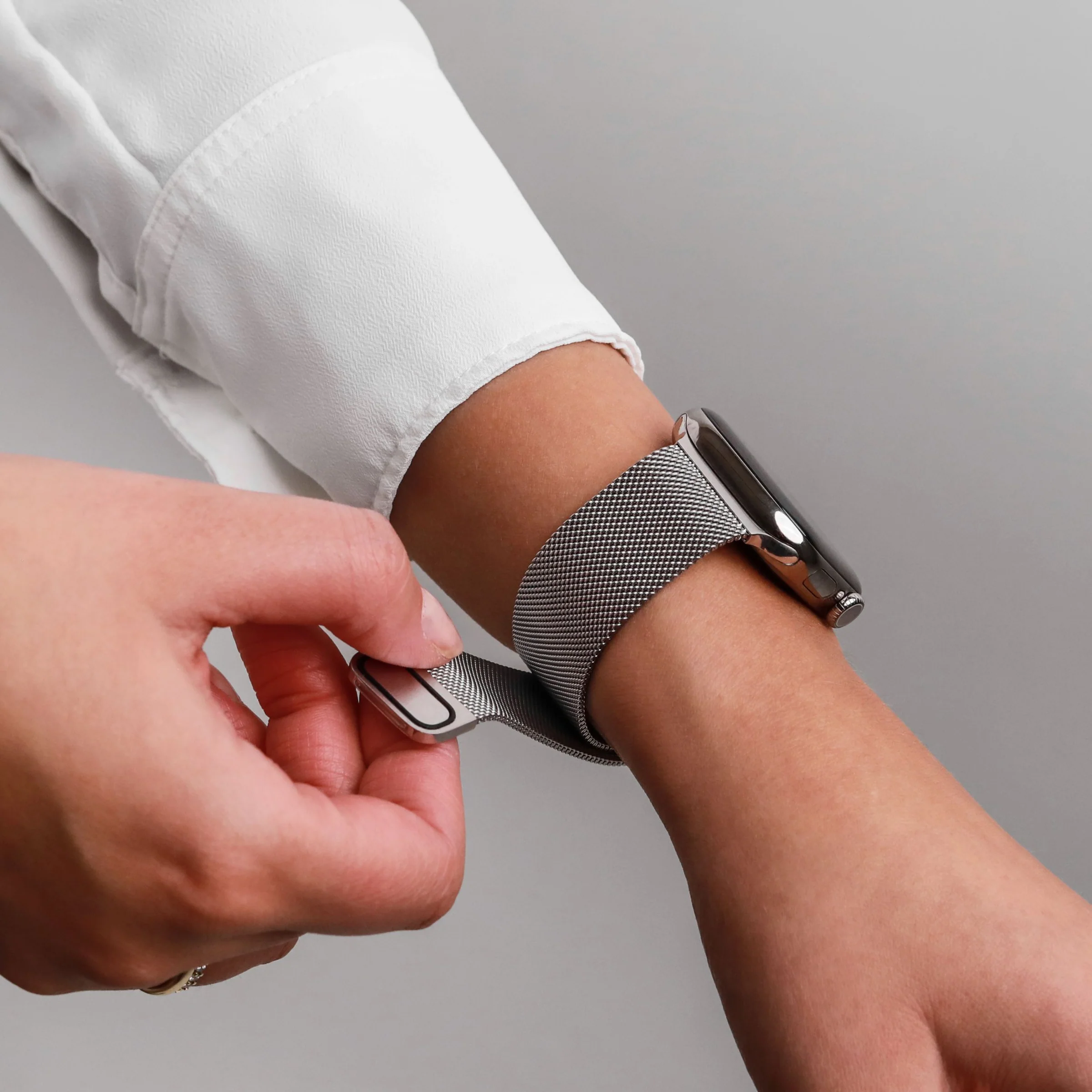 Une main attache le bracelet argenté d'une montre connectée à une chemise blanche.