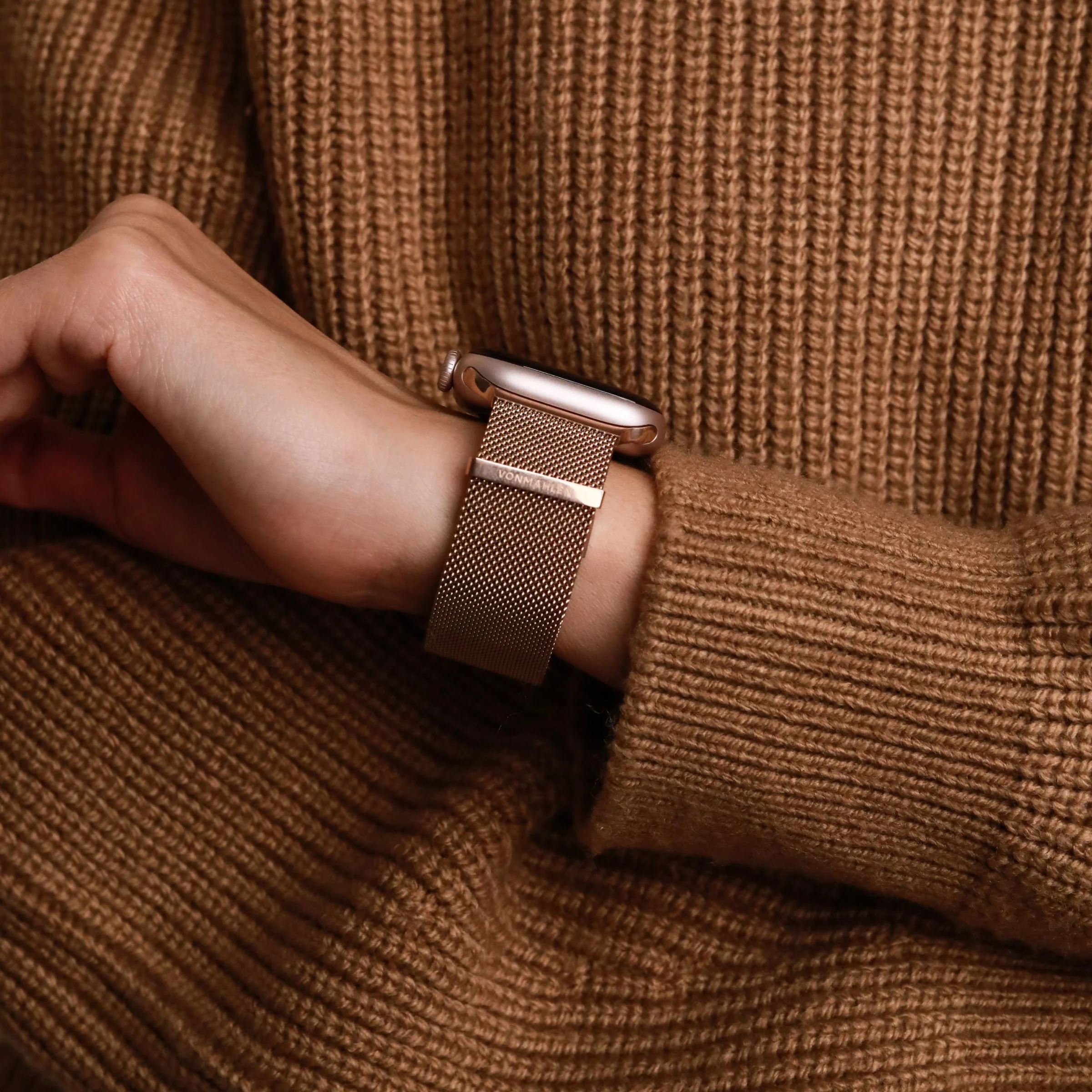 Le poignet d'une personne porte une montre connectée or rose avec un bracelet en maille, sur un pull en tricot marron.