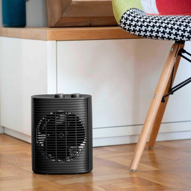 Un calefactor eléctrico portátil negro se encuentra sobre un suelo de madera junto a una silla.