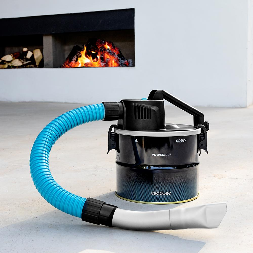 Un aspirador de cenizas negro con manguera azul ante una chimenea con leña ardiendo.