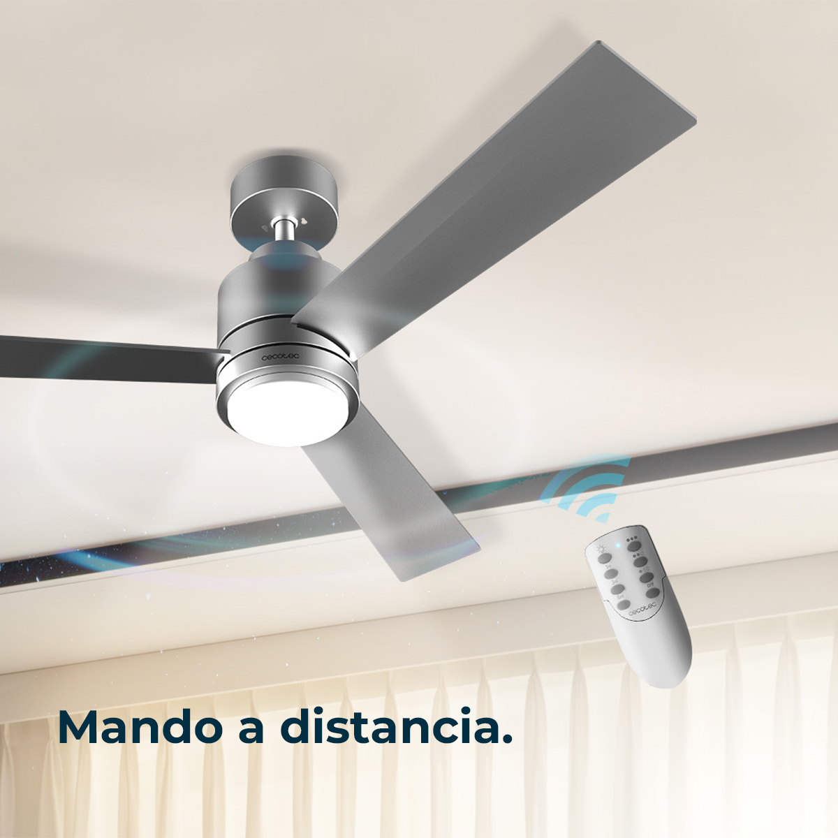 Un ventilador de techo plateado con mando a distancia frente a cortinas transparentes.
