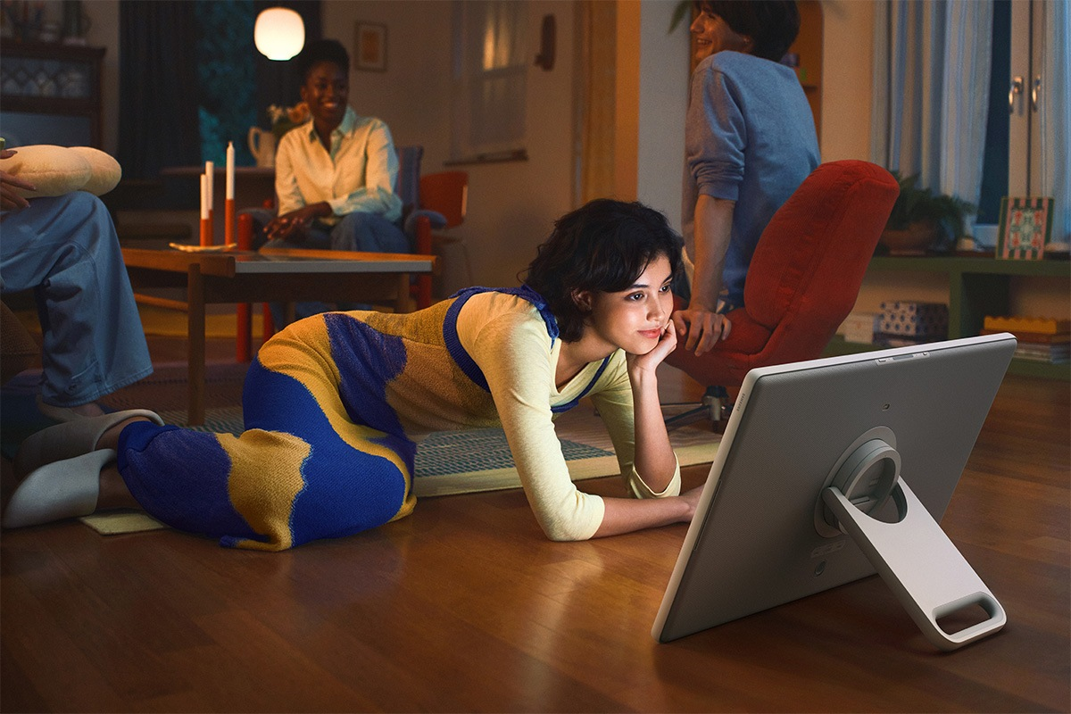 Una donna in abito blu e giallo guarda un tablet sul pavimento. Altri sono seduti sullo sfondo.