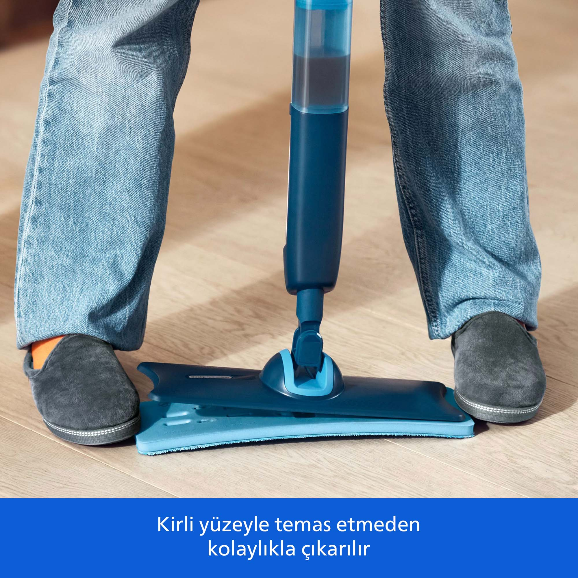 Kot pantolonlu ve gri terlikli kişi ahşap zeminde mavi sprey paspas kullanıyor.