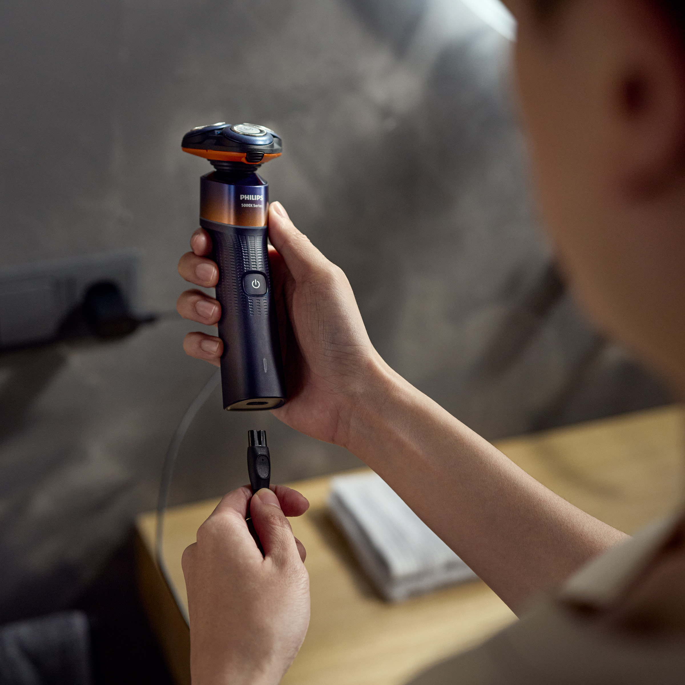 Dłoń trzyma ciemną maszynkę do golenia z pomarańczowymi akcentami, podłączając kabel ładujący.