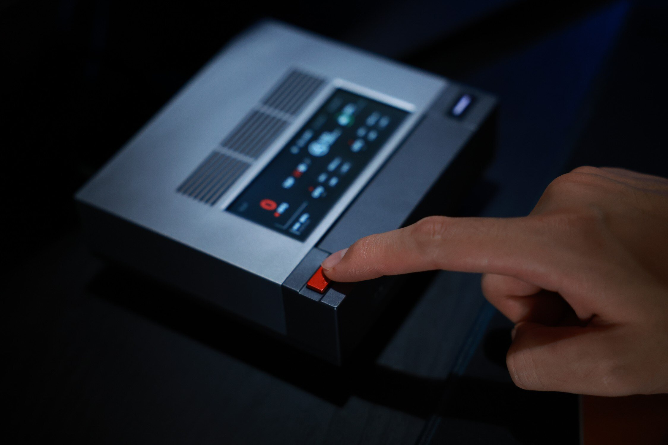 Dłoń naciska czerwony przycisk na szarym urządzeniu z ekranem.