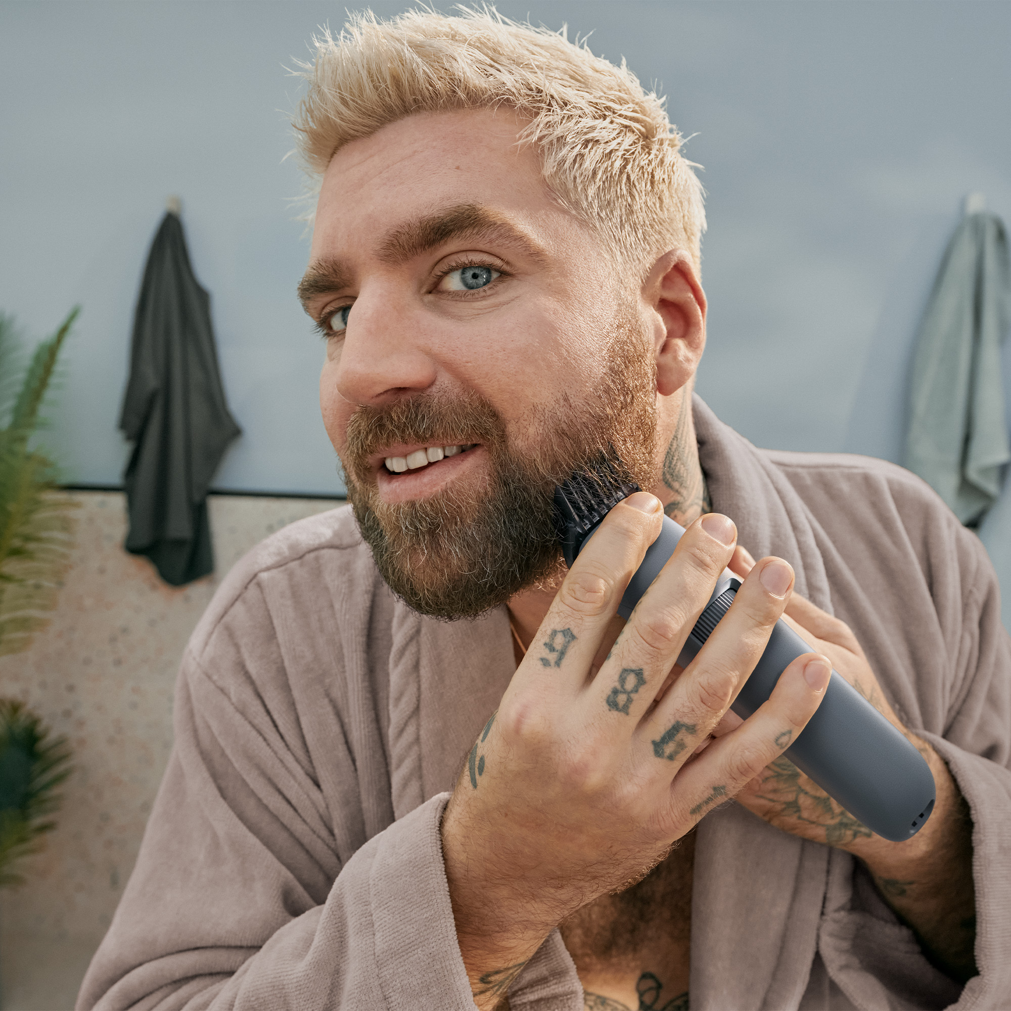 Uśmiechnięty mężczyzna z blond włosami i niebieskimi oczami, w brązowym szlafroku, przycina brodę szarą golarką elektryczną.