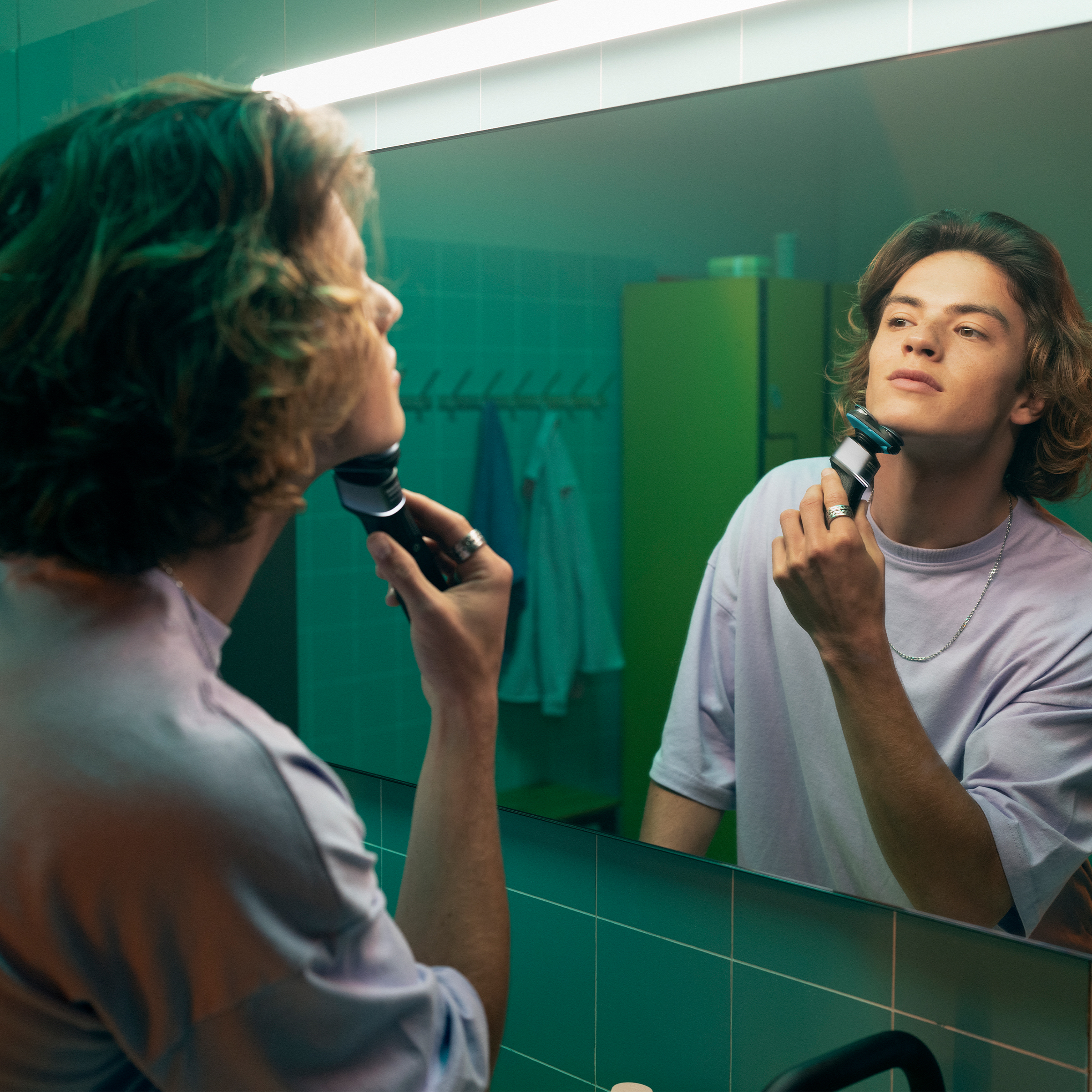 Młody mężczyzna goli twarz w lustrze golarką elektryczną.