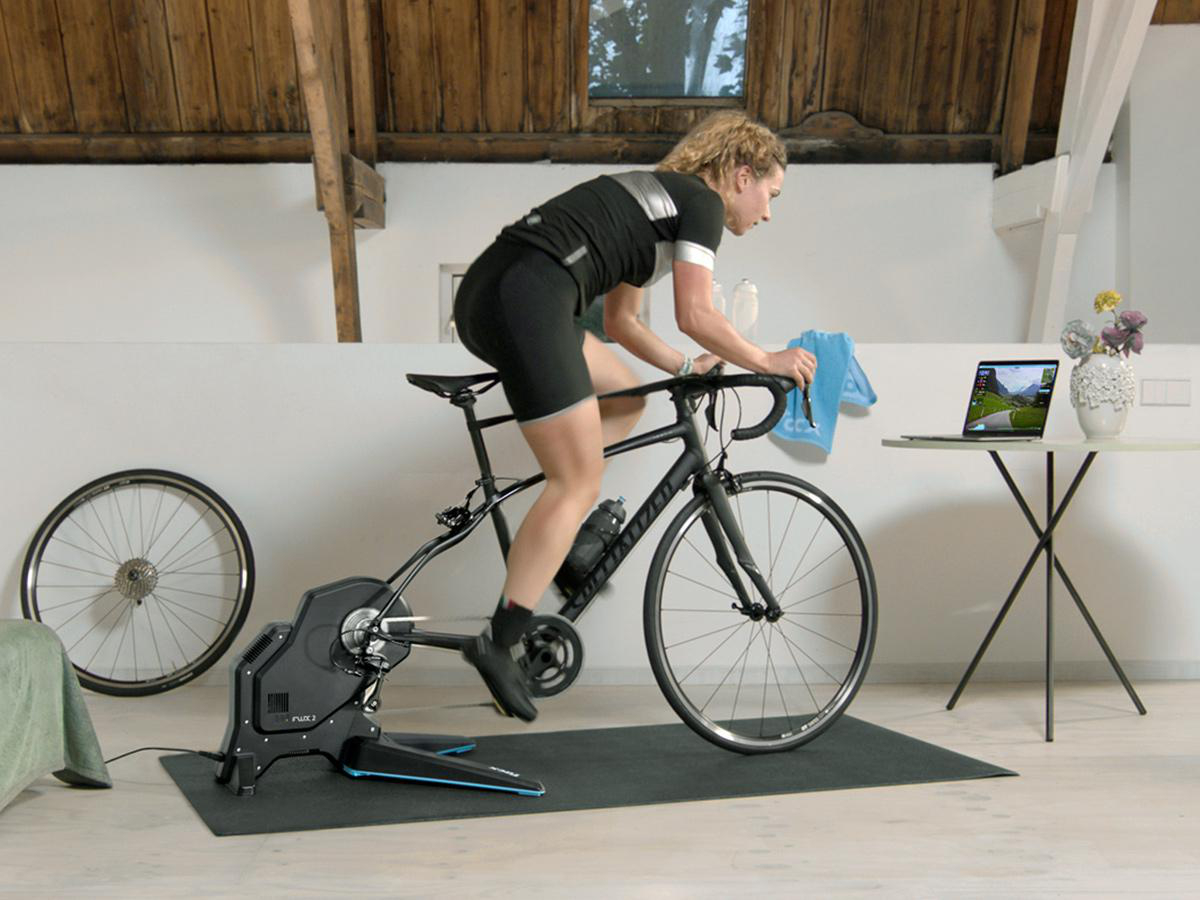 Frau auf einem Heimtrainer fährt Rad, schaut auf einen Laptop mit virtueller Landschaft.