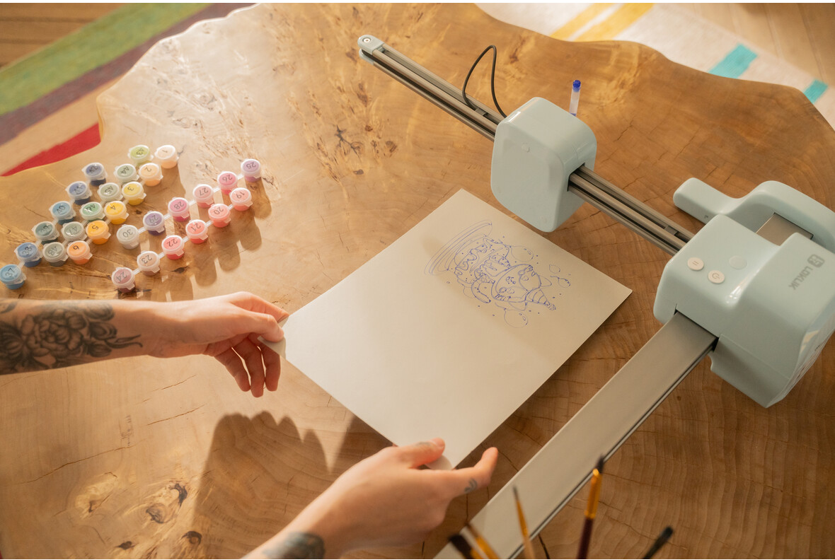 Un robot da disegno su tavolo di legno, con vasetti di vernice e carta.