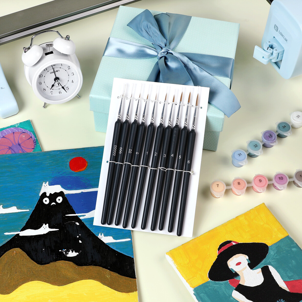 Materiali artistici: pennelli, vasetti di vernice, scatola regalo, sveglia e dipinti.