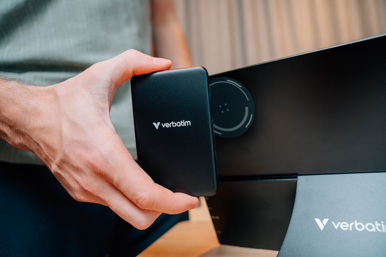 Una mano tiene un hard disk Verbatim nero accanto a uno schermo nero.