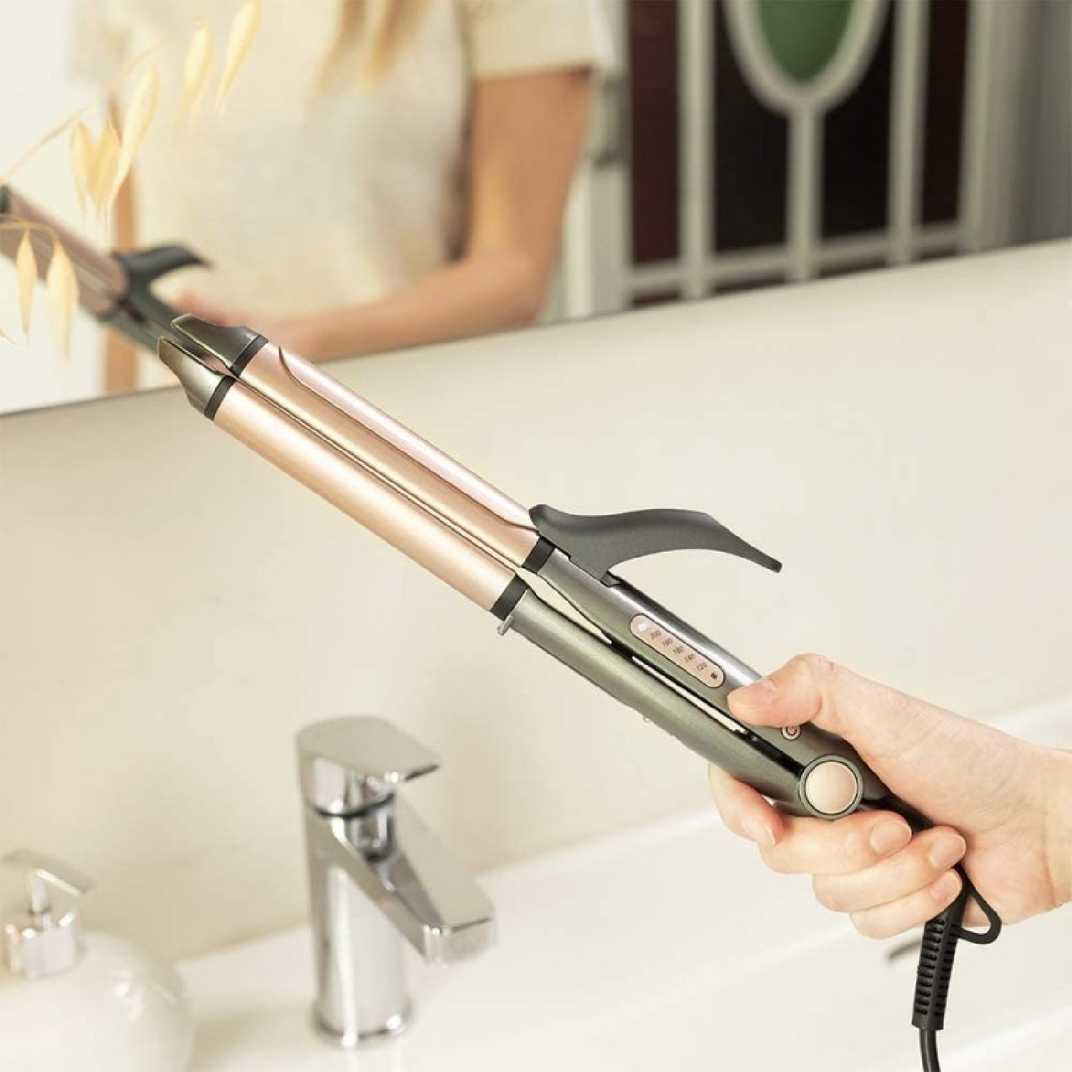 Una mano sostiene un rizador oro rosa y gris frente a un lavabo.