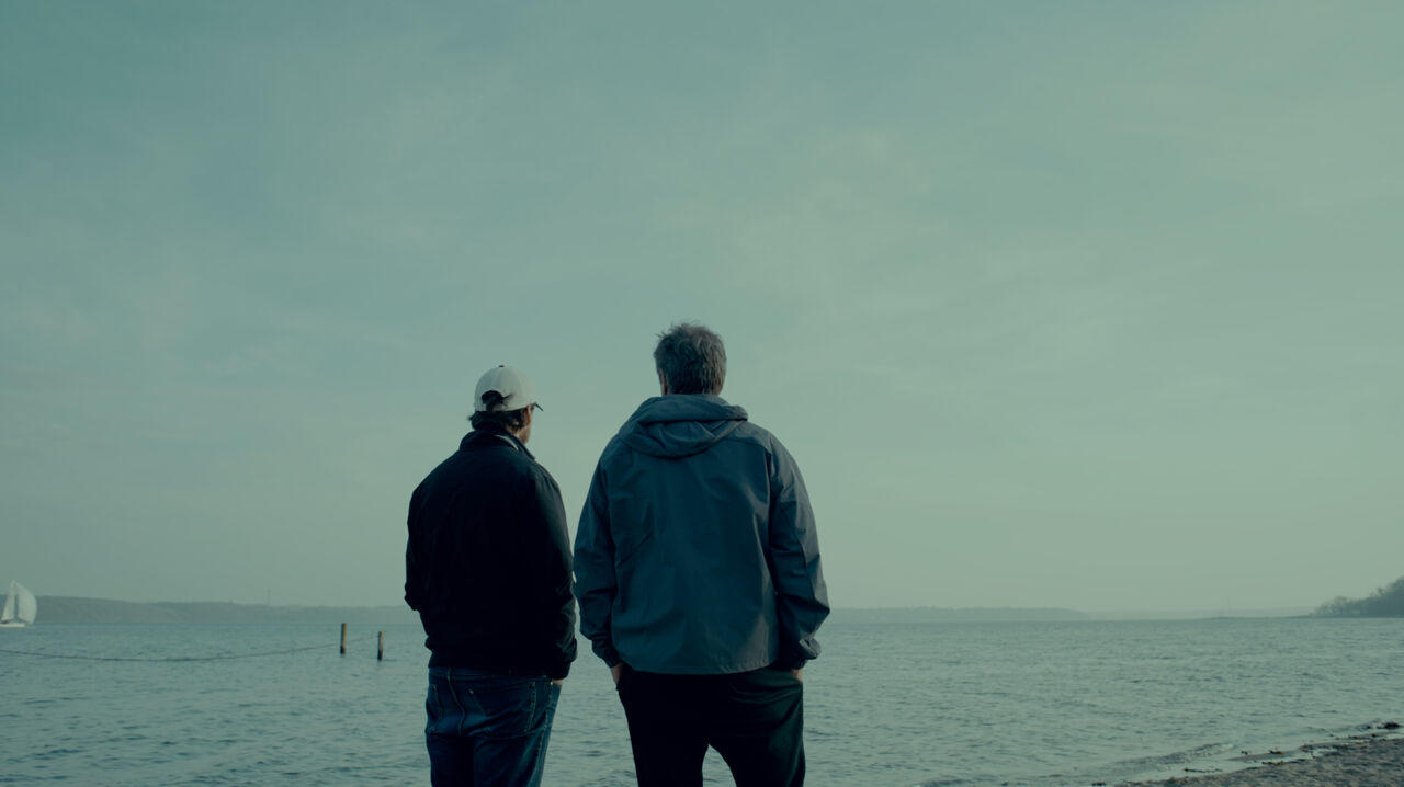 Zwei Männer stehen am Ufer, schauen aufs Meer. Ein Segelboot ist links.