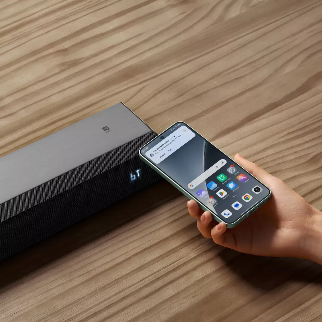 Une main tient un téléphone se connectant à une barre de son grise sur une table en bois.