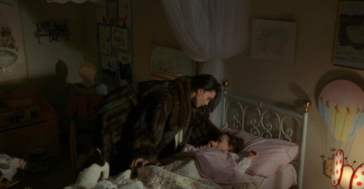 Eine Frau in einem dunklen Pelzmantel beugt sich über ein kleines Kind, das in einem schwach beleuchteten Zimmer in einem Bett schläft.