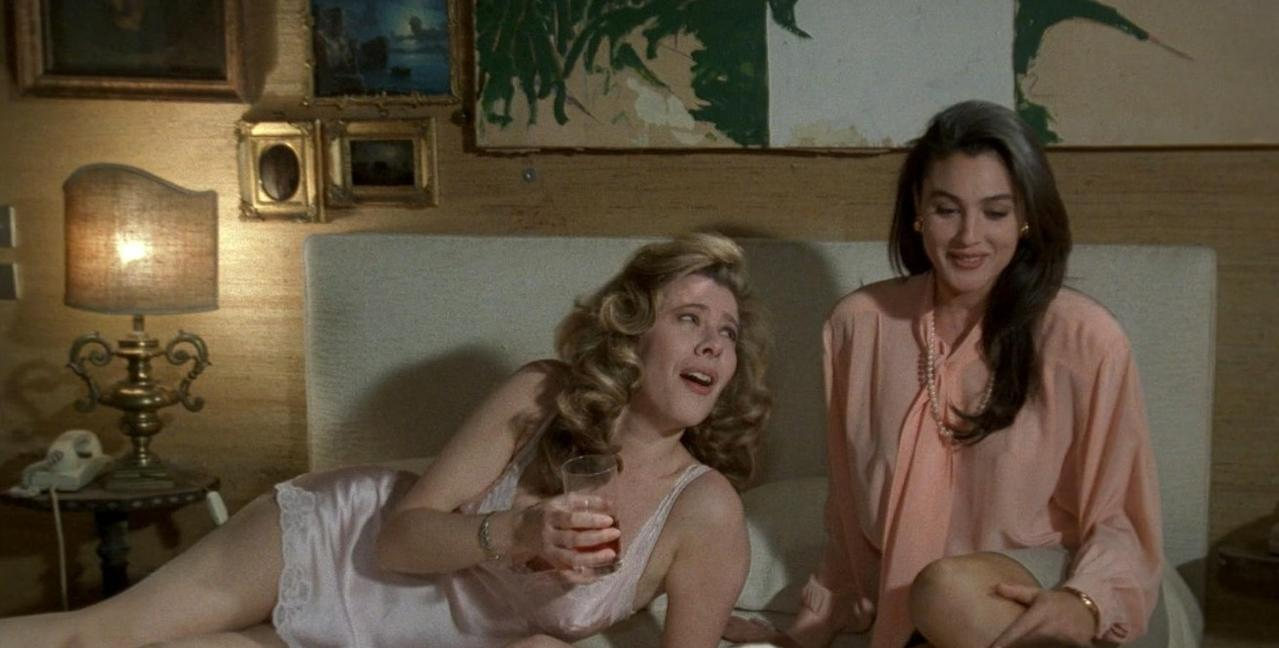 Zwei Frauen sitzen auf einem Bett. Eine hält ein Glas, die andere trägt eine rosa Bluse.