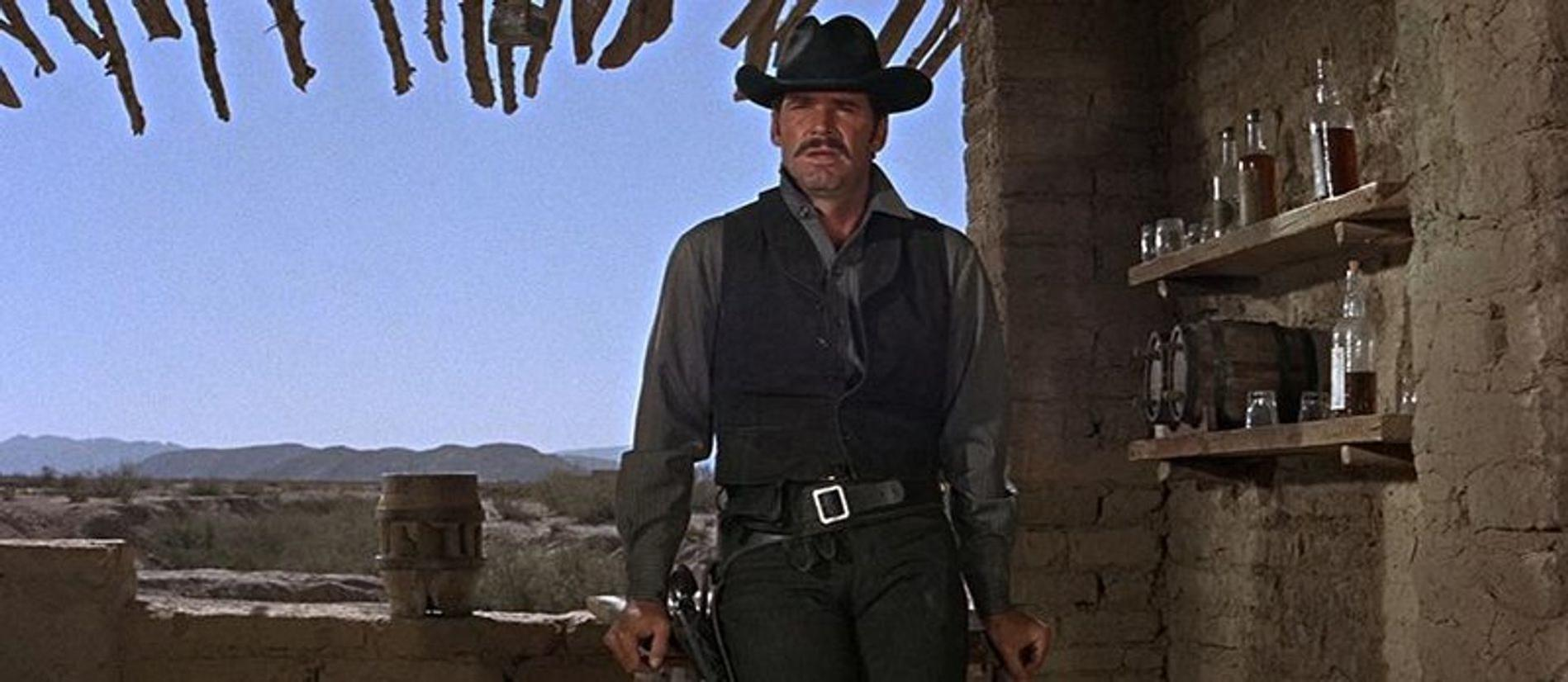 Ein Mann im Western-Look steht vor einer Bar, im Hintergrund Wüste.