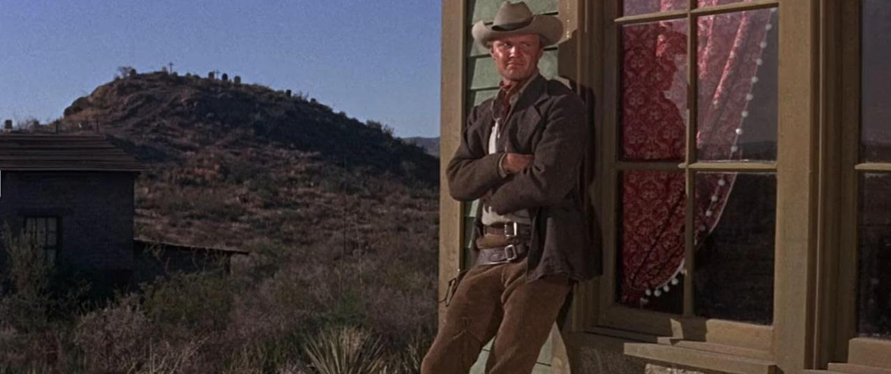 Ein Cowboy mit Hut und Mantel lehnt an einem Fenster. Im Hintergrund Hügel mit Gräbern.