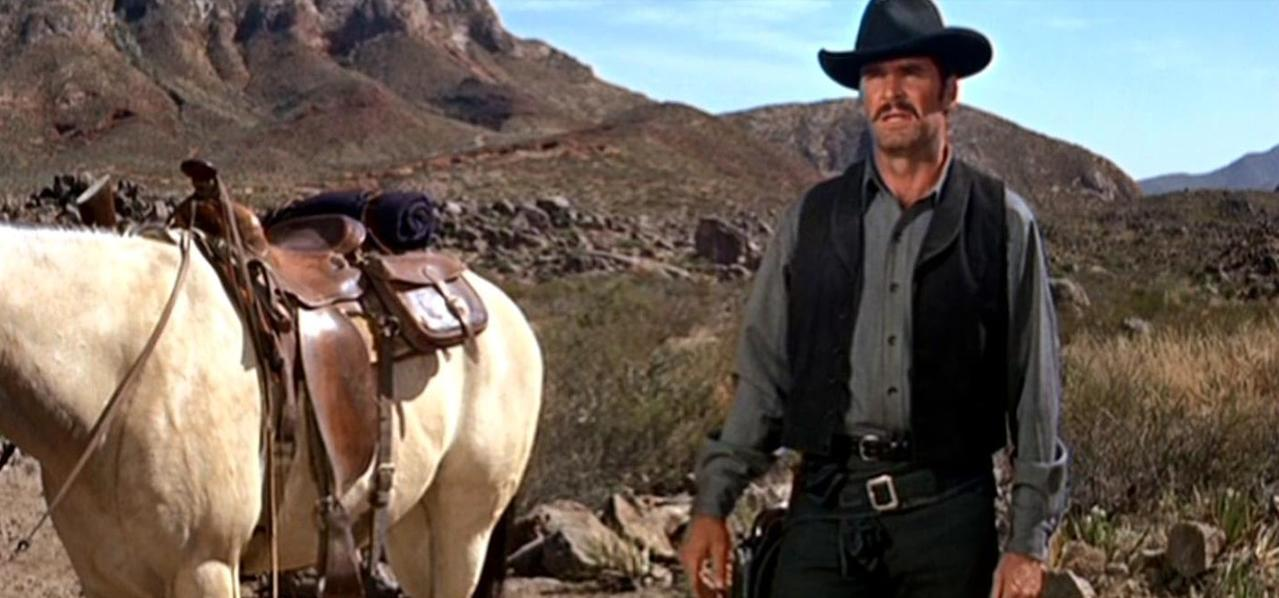 Ein Cowboy in schwarzem Hut und Weste steht neben einem hellen Pferd mit Sattel.