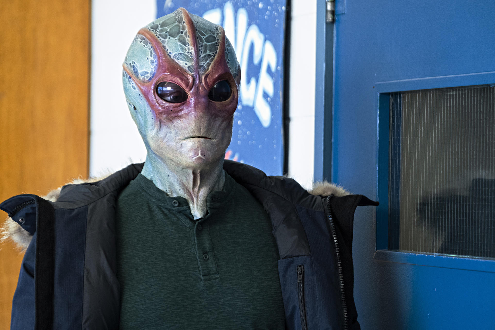Ein Alien mit blau-grüner Haut, roten Akzenten und dunklen Augen trägt einen grünen Pullover und eine Jacke.