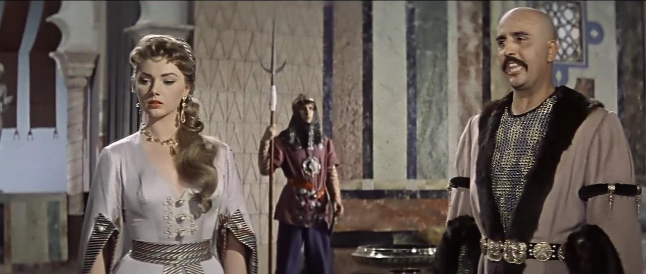 Eine Frau in einem Kleid steht links, ein Mann mit Glatze rechts, ein Soldat im Hintergrund.