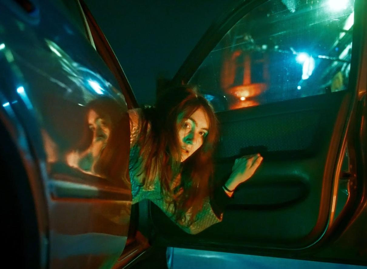 Eine Frau mit braunen Haaren blickt aus einem Auto bei Nacht, umgeben von grünem Licht.