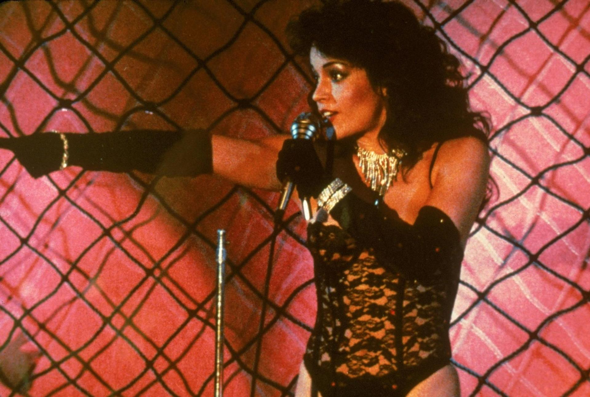 Eine Frau mit Mikrofon und Glitzerkette singt vor einem Netz mit rosa Hintergrund.