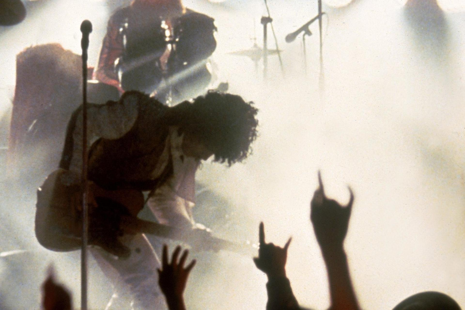 Ein Gitarrist spielt auf einer Bühne im Nebel, Fans machen Rock-Geste.