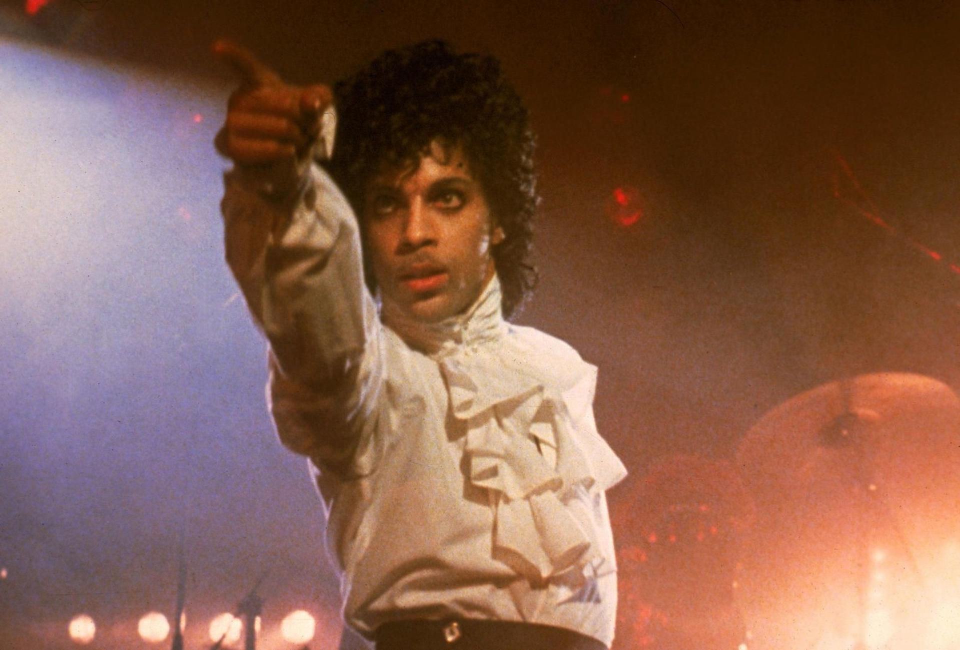 Prince in weißer Rüschenbluse zeigt auf die Kamera, auf dunkler Bühne.