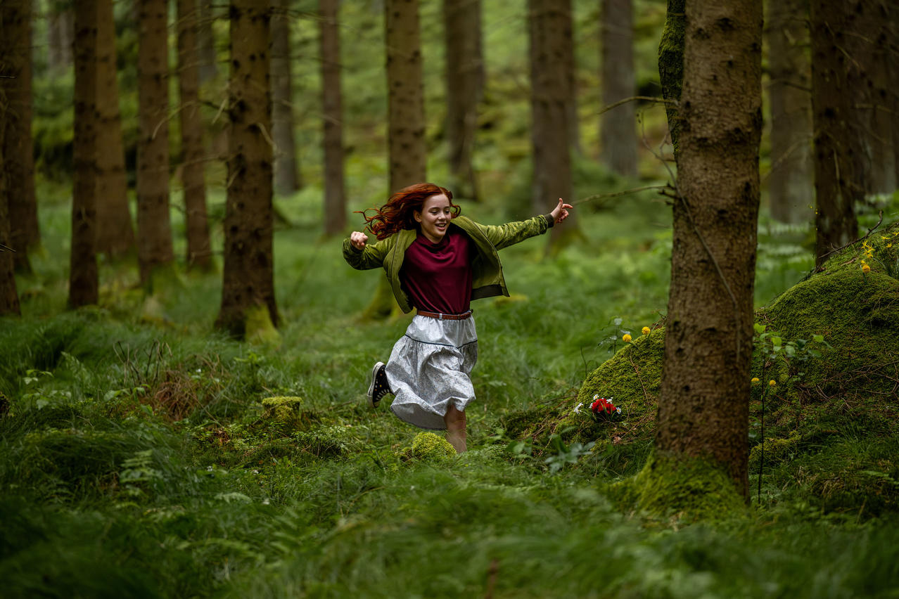 Ein Mädchen mit roten Haaren rennt lachend durch einen grünen Wald.