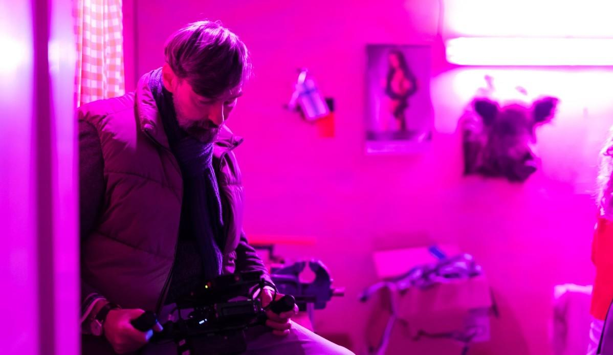 Mann mit Kamera vor rosa Wand mit Kuhkopf und Poster.
